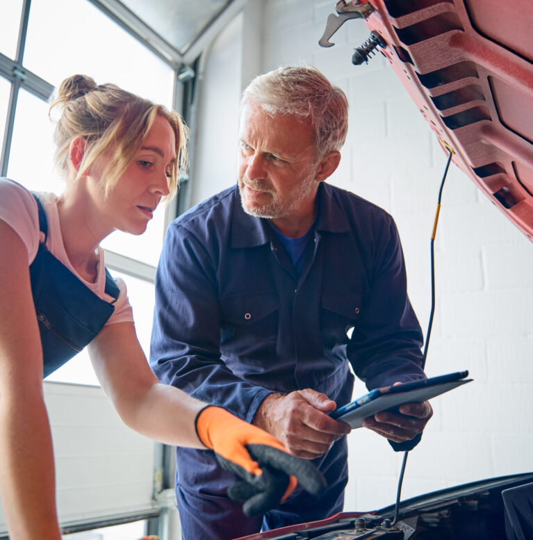 Male Car Auto Mechanic With Female Trainee Looking Under Bonnet Of Car Holding Digital Tablet